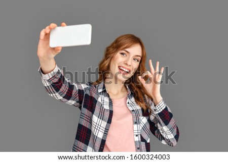 Young stylish woman studio standing isolated on grey background taking selfie photo on smartphone showing ok sign to camera smiling playful