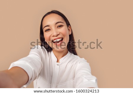 Young woman wearing white shirt with dark long hair standing isolated on bage background taking selfie photo on smartphone looking camera laughing happy close-up