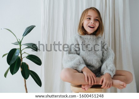 Happy little barefoot girl in a grey knitted sweater sitting cross-legged on a stool at home, in front of a curtain next to a potted plant.