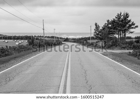 A black and white perspective photo of a long country road stretching to the Atlantic coast between farm fields under storm clouds. 