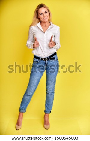 Full-length art portrait of a pretty smiling blonde woman in blue jeans and a white shirt stands on a yellow background. Right in front of the camera with emotions.