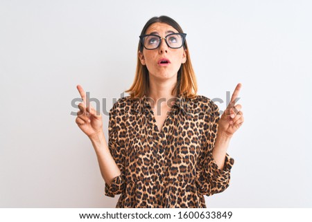 Beautiful redhead woman wearing glasses and elegant animal print shirt over isolated background amazed and surprised looking up and pointing with fingers and raised arms.