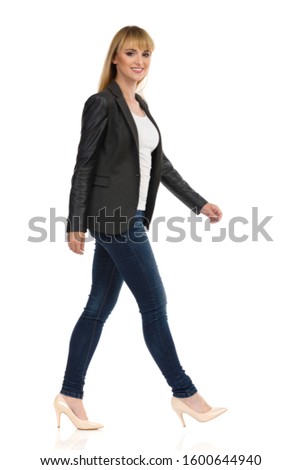 Smiling young businesswoman in black suit, blue jeans and beige high heels is walking and looking at camera. Full length studio shot isolated on white background.