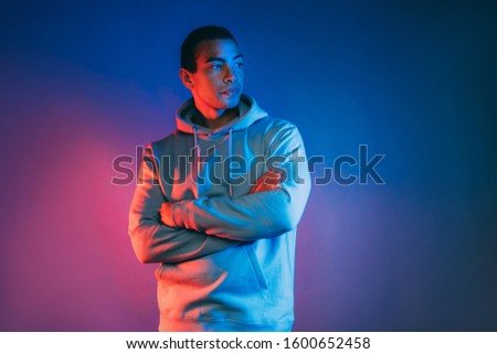 Handsome cool attractive young arabian man on picture. Side view of strong guy holding hands crossed and look to right. Wear blue hoodie. Isolated over multicolored background
