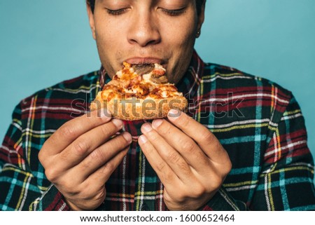 Close up and cut view of young arabian or egyptian man posing hold slice of pizza with hands and bite it. Guy in checkered shirt enjoy moment. Eating fat unhealthy meal. Isolated over blue background