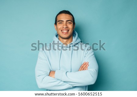 Young happy latin arabian or hispanic man posing on camera and smile. Hold hands crossed. Guy wear blue stylish hoodie. Stand alone and rejoice. Isolated over blue background