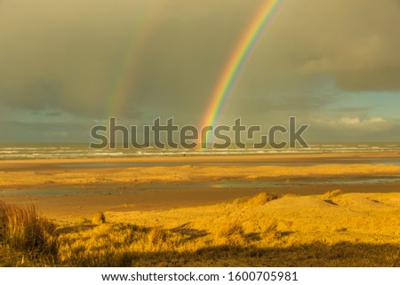 Sunrise with a view over the Dutch North Sea beach and dunes near IJmuiden towards the sea with bright harsh sunlight against rain clouds with two rainbows above the sea