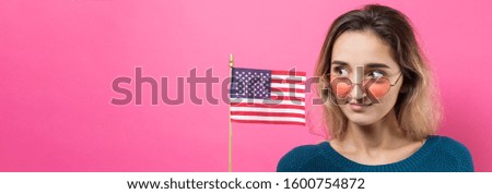 Happy young woman heart-shaped glasses holding American flag against a studio pink background