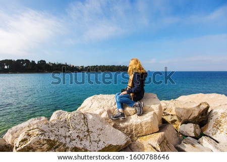 Back view of a girl. Woman sitting on the rocky shore of the sea. Looking away. Looking straight ahead. Rear view of a lonely young adult person. A beautiful seaside landscape. Blue sky. 
Europe