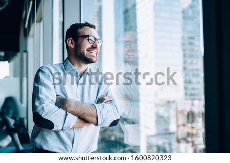 Cheerful male entrepreneur with crossed hands standing near office window view and smiling during work day in company, Caucasian successful corporate boss feeling good from wealthy lifestyle Royalty-Free Stock Photo #1600820323