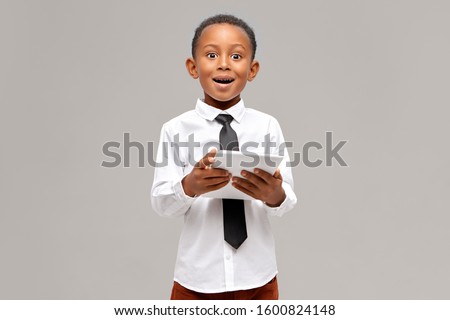 Astonished dark-skinned little boy wearing white shirt and black tie enjoying high speed wireless internet connection on digital tablet having surprised amazed look, watching cartoon online