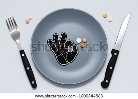 Multicolored pills and okay symbol on a gray plate next to a knife and fork on a white background. Medical concept.