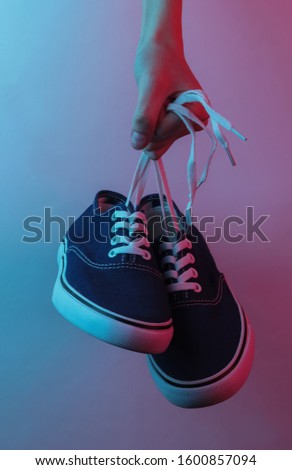 Female hand holding sneakers by the laces. Creative pop art pink blue neon color. Trendy gradient illumination. Night light