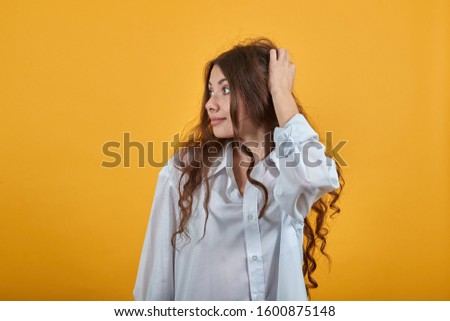 Curly caucasian young woman in fashion white shirt keeping hands on head, looking aside, having nice haircut isolated on orange background in studio. People sincere emotions, lifestyle concept.