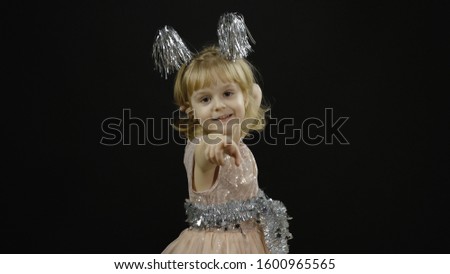 Happy beautiful little baby girl in glossy dress emotionally points at something with her hands. Christmas concept. Positive, pretty, four years old child make faces and smile. Black background