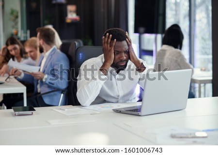 Worried afro-american office manager disappointed looking at laptop screen in the office room on the background of his coworkers. Business issues concept.