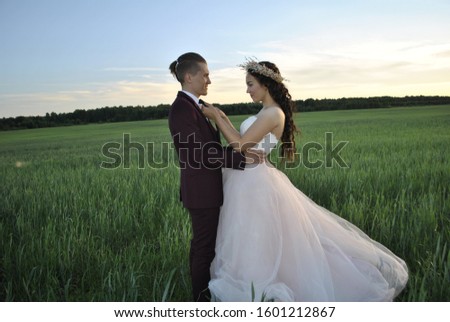 Wedding couple at a photo shoot in a wheat field. The bride straightens the groom's tie. The groom holds the bride by the waist. The young couple smiles.