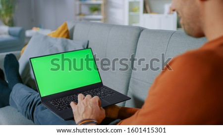 Man Lying on His Sofa At Home, Uses Laptop with Green Mock-up Screen. Over The Shoulder Shot of Anonymous Man in the Living Room.