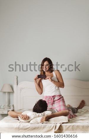 Young cheerful woman using cell phone and taking picture of her boyfriend in bed