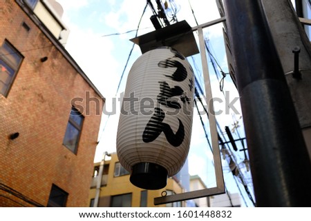Text on lantern is “udon” which means udon noodles. Unlit lantern advertising udon noodles restaurant in Japanese hiragana.  Street sky view backdrop in background in Japan with electrical wires.