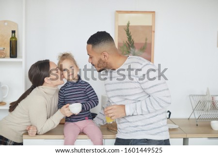 Portrait of happy mixed-race family kissing cute little girl while enjoying breakfast together in modern kitchen interior, copy space