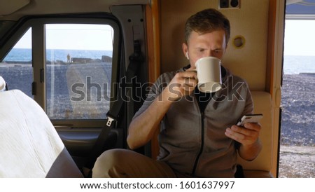 man drinks coffee and uses a smartphone sitting in a caravan by the sea. Royalty-Free Stock Photo #1601637997