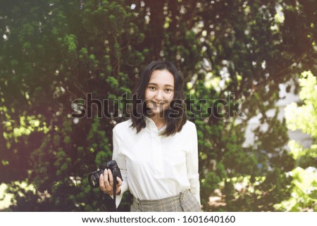 Optimistic asian girl in white shirt in a park, smiling, holding camera. Outdoor portrait.