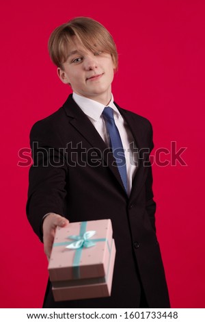 Young teenager guy in a black jacket holds a box with a gift on a red background