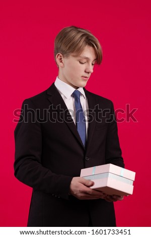Young teenager guy in a black jacket holds a box with a gift on a red background
