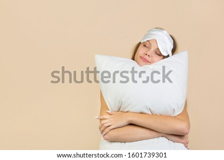 beautiful young woman on a beige background in a sleep mask with a pillow in her hands Royalty-Free Stock Photo #1601739301