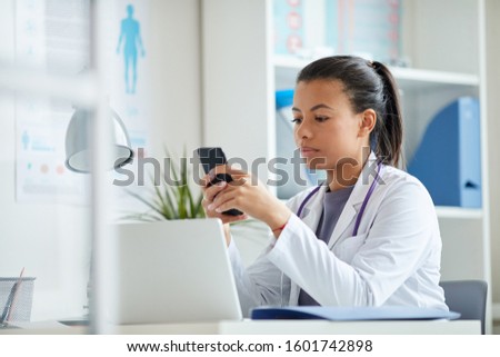 Young female doctor sitting at the table and using her mobile phone during her work at office