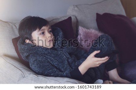 Cute kid sitting on sofa holding remote control, A happy child boy relaxing at home watching TV during cold weather outside in Autumn or Winter. Indoors activities for kids during bad weather 