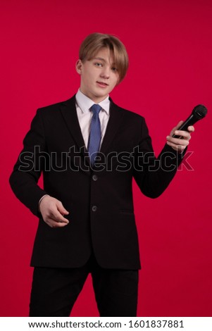 Teen boy in a black jacket holds a microphone in his hands and poses on a red background