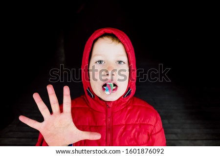 Scary boy with red hood lit with neons is a strange pose while eating a candy.