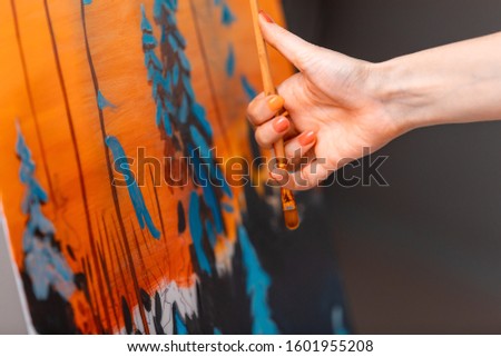 The artist’s hand measures perspective while painting the winter forest at sunset in orange and blue tones in studio. Christmas and New Year concept, arts. 