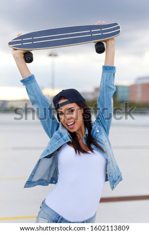 Happy young urban woman holding longboard over head