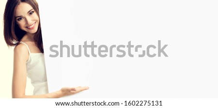 Portrait of happy smiling young woman in white casual smart clothing, showing empty blank signboard with copyspace area for text or slogan, isolated over yellow background
