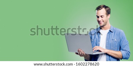 Smiling attractive man in blue smart casual wear, working with laptop, with empty copy space area for some slogan, advertisement or text, against green background. Banner composition.
