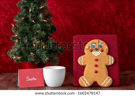 Large gingerbread cookie on a red background, Christmas tree with white lights, note to Santa, white mug with milk, wood table
