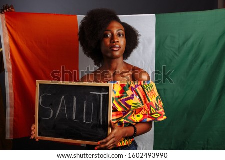 Portrait of young African woman student in national clothes holding a chalkboard with Hello word on her native language before the Ivory Coast flag..