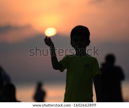 Silhouette image of young boy standing and showing the visuals like he is holding sun at sunset on shore of beach in winter
