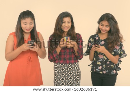 Studio shot of three happy young Persian woman friends smiling while using mobile phones together against white background
