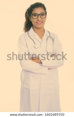 Studio shot of young happy Persian woman doctor smiling and standing while wearing eyeglasses with arms crossed isolated against white background