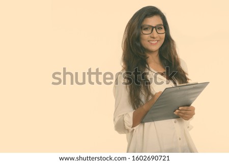 Studio shot of young happy Persian woman smiling while holding clipboard with eyeglasses isolated against white background