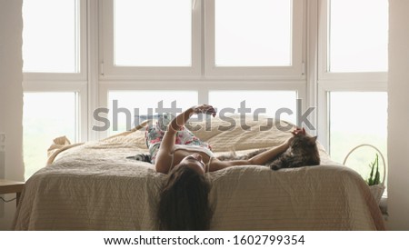 Young beautiful woman relaxing with her lovely Maine Coon cats laying in bed uses mobile phone by the window