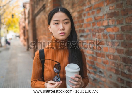 Portrait of young Asian woman holding a cup of coffee while standing outdoors in the street. Urban concept.