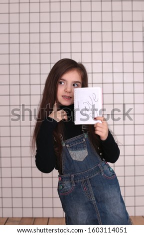 young smiling girl of eight years with long hair in a black turtleneck and denim sundress with a sign "love and heart"
