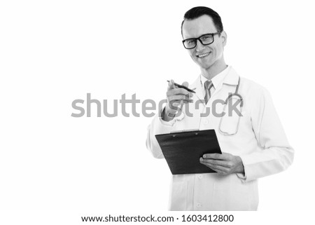 Studio shot of young happy man doctor smiling while holding clipboard and pen while pointing at camera