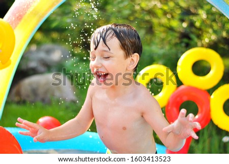 Laughing boy under water drops shower. Sunny summer mood picture. Cheerful kid playing in swimming pool with water drops under the bright sunshines. Childish playful joy and happy childhood concept.