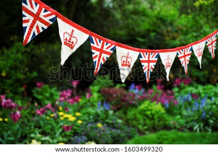 Banner of British Union Jack flag and royal crown celebratory bunting hanging in front of a bright English summer garden background Royalty-Free Stock Photo #1603499320